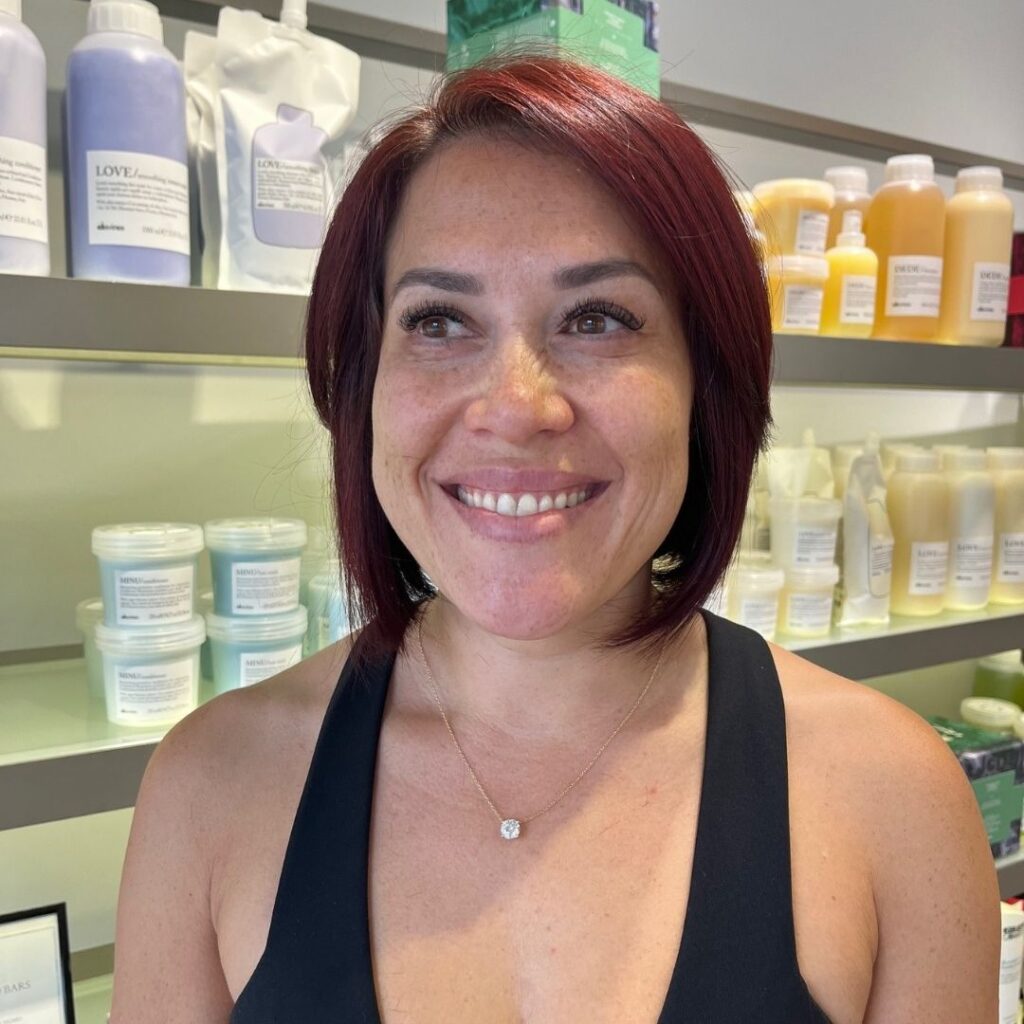 Person with short red hair smiling in a store with shelves of skincare products in the background. - Detlev Lifestyle Salon in Coconut Grove, FL