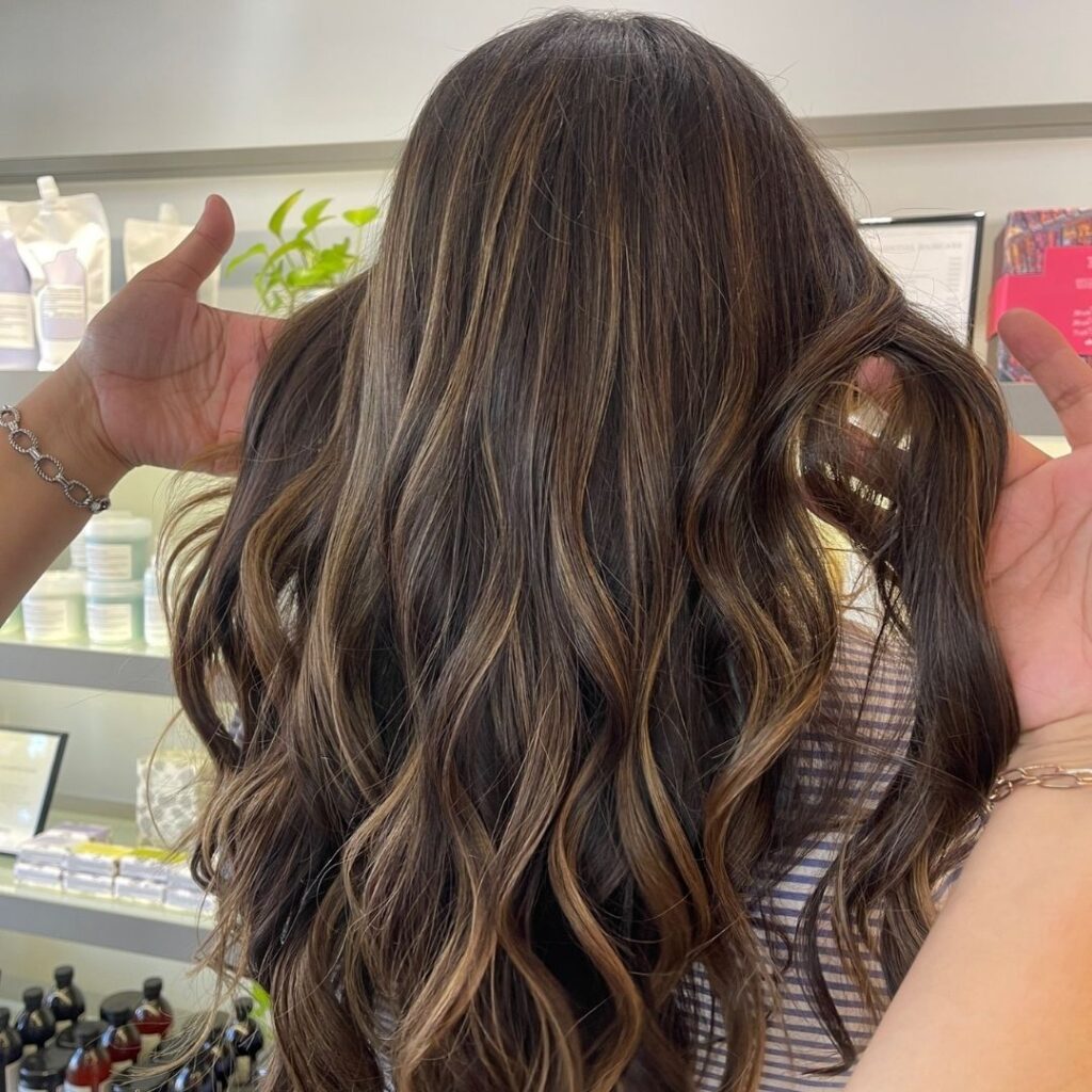 Person with long, wavy, brown hair with highlights, shown from the back. A hand on the sides holding the hair. Shelves with products in the background. - Detlev Lifestyle Salon in Coconut Grove, FL