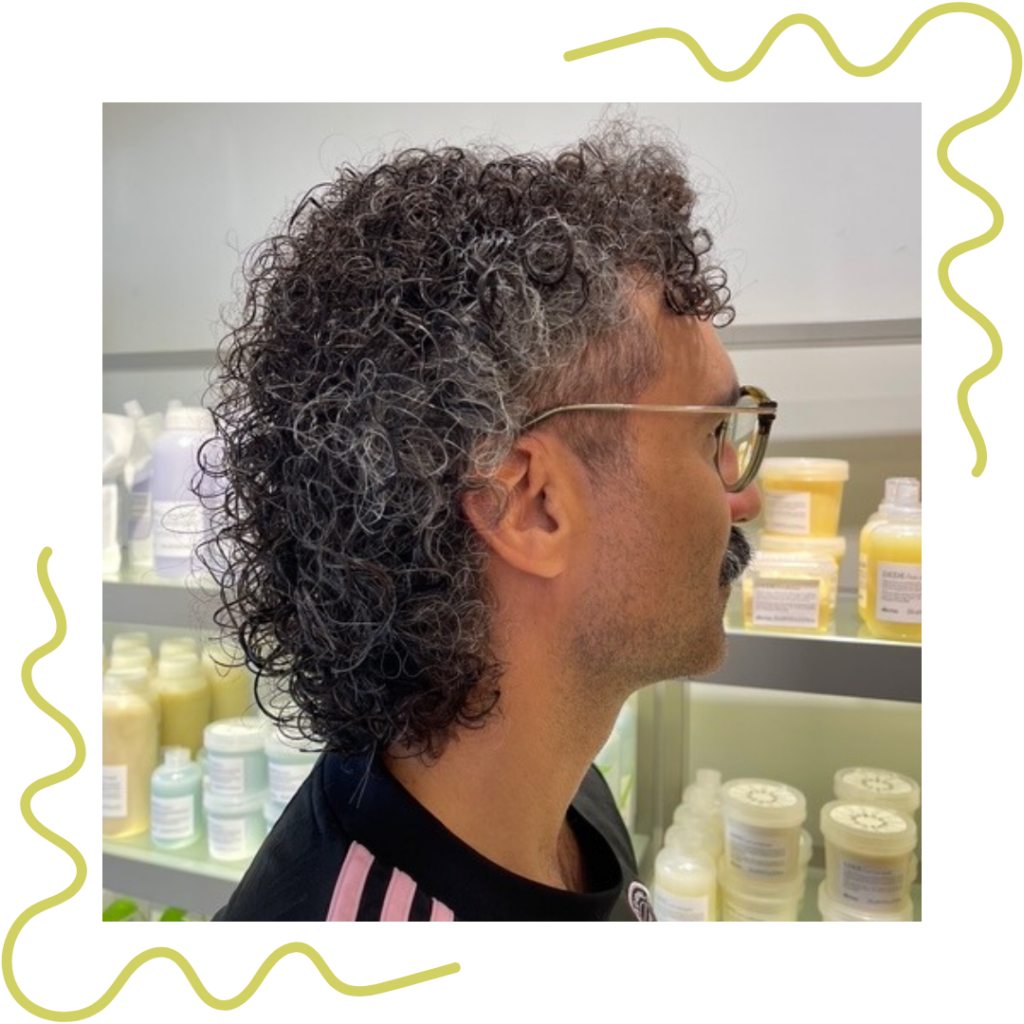 Person with curly, shoulder-length hair and glasses stands in profile. Shelves of hair products are visible in the background. - Detlev Lifestyle Salon in Coconut Grove, FL