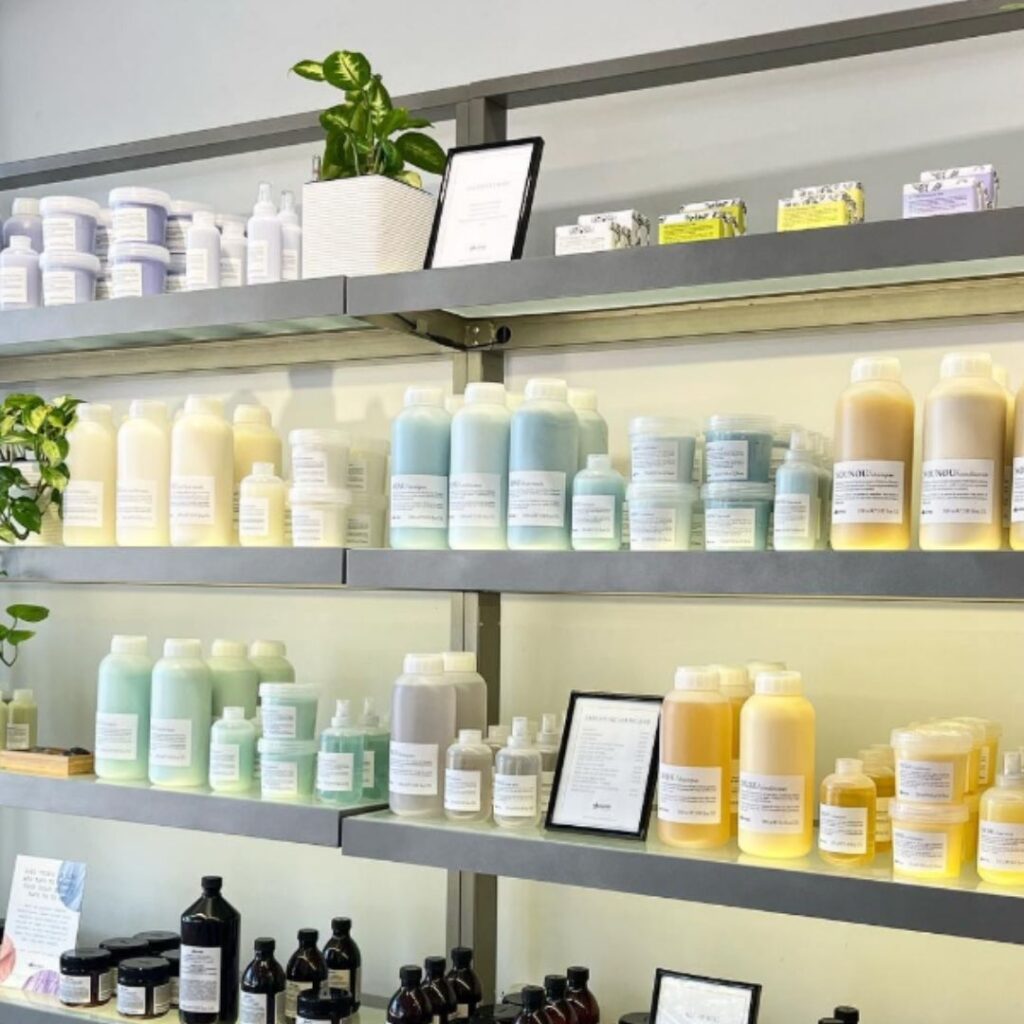 Shelves display various bottles and containers of skincare products, arranged by color and size. Greenery and small plaques are placed alongside. - Detlev Lifestyle Salon in Coconut Grove, FL