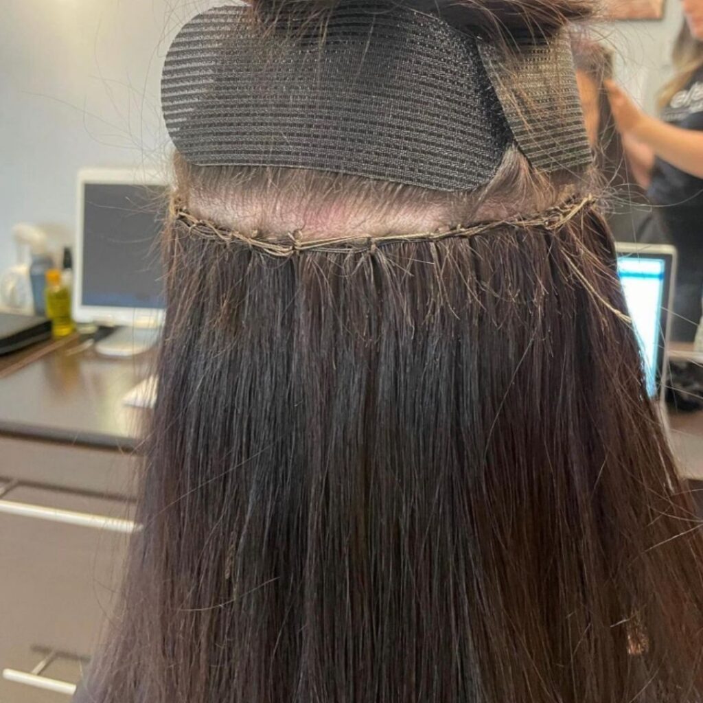 Close-up of hair extensions being applied to a person's head, with a section clipped up. Monitors and a stylist are visible in the background. - Detlev Lifestyle Salon in Coconut Grove, FL