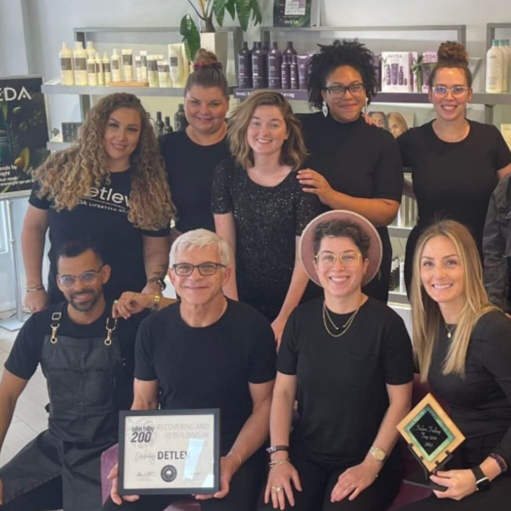 A group of nine people, some seated and some standing, pose together in a hair salon. Two individuals hold plaques, and hair products are visible in the background. - Detlev Lifestyle Salon in Coconut Grove, FL