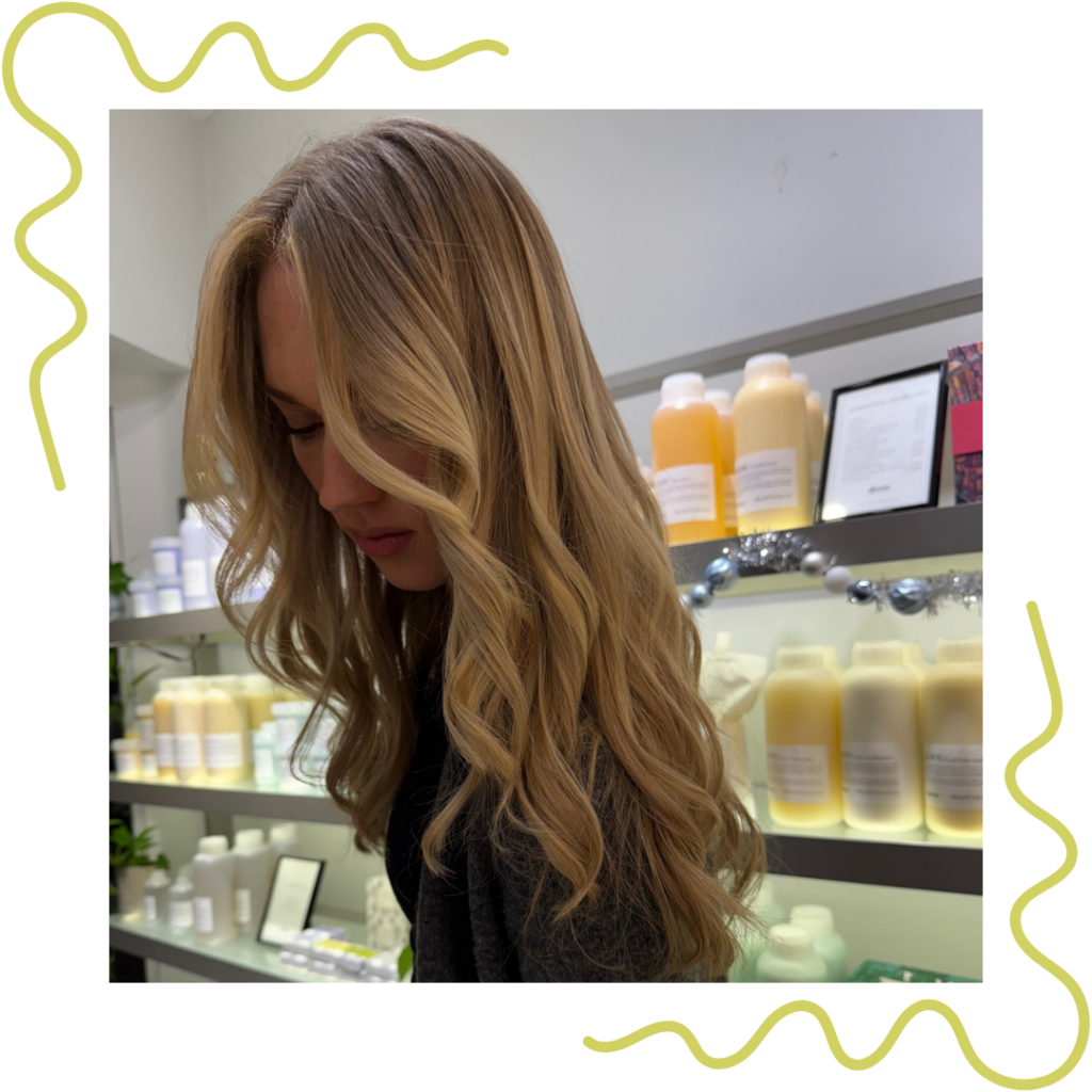 Person with long, wavy blonde hair in a salon with shelves displaying various hair products. - Detlev Lifestyle Salon in Coconut Grove, FL