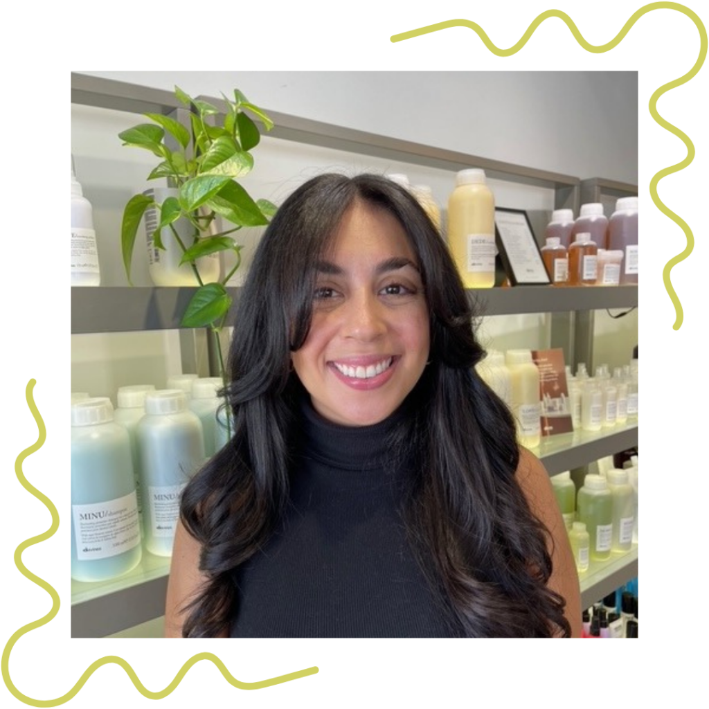 Woman with long dark hair smiles in front of shelves filled with various bottled products. - Detlev Lifestyle Salon in Coconut Grove, FL