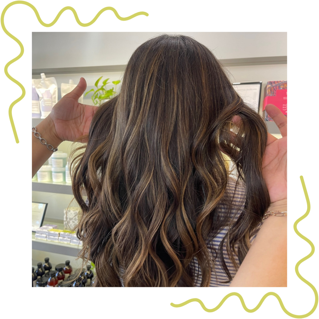 A person is showing the back of their wavy, dark brown hair with highlights in a salon. Hands are holding sections of the hair. Various products are visible in the background. - Detlev Lifestyle Salon in Coconut Grove, FL