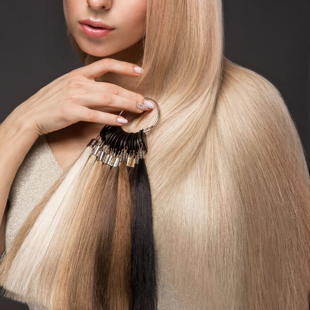 A woman with long, straight blond hair showcases a ring of hair color samples against a dark background, highlighting her healthy hair even in humid climates. - Detlev Lifestyle Salon in Coconut Grove, FL