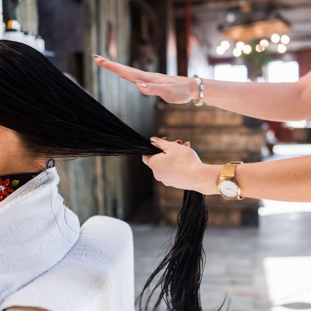 A person with wet hair sits in a salon chair, focusing on scalp health, while another gently combs through their long, black locks using fingers and palm. - Detlev Lifestyle Salon in Coconut Grove, FL