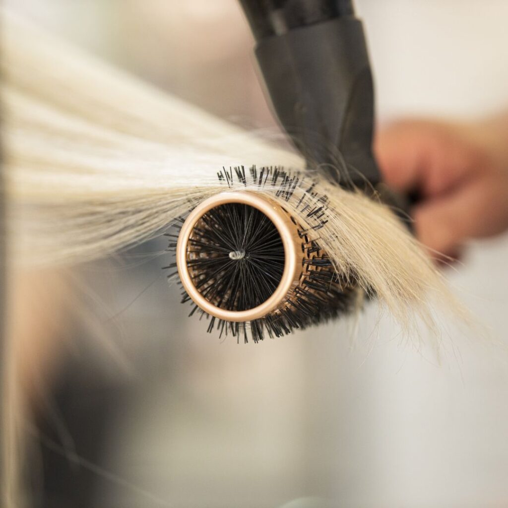 Close-up of a round brush and blow dryer being used to style straight blonde hair, showcasing the smooth finish achieved with professional hair extensions. - Detlev Lifestyle Salon in Coconut Grove, FL
