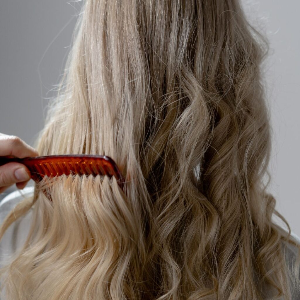A person with long, wavy blonde hair—styled with Bellami Hair Extensions—is being combed from behind using a wide-tooth, red comb. - Detlev Lifestyle Salon in Coconut Grove, FL