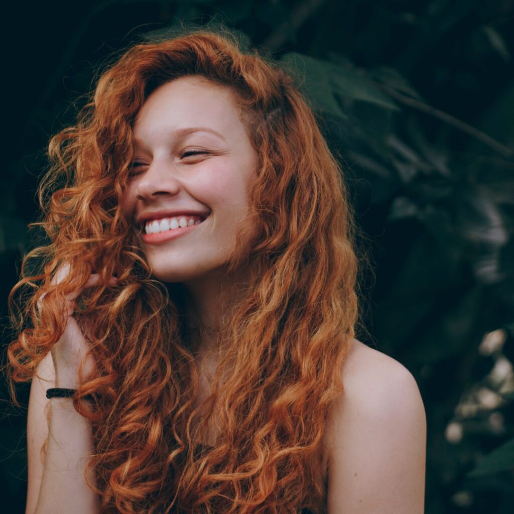 A woman with long, curly red hair smiles and looks to the side, standing in front of leafy green foliage, showcasing beautiful Bellami Hair Extensions. - Detlev Lifestyle Salon in Coconut Grove, FL