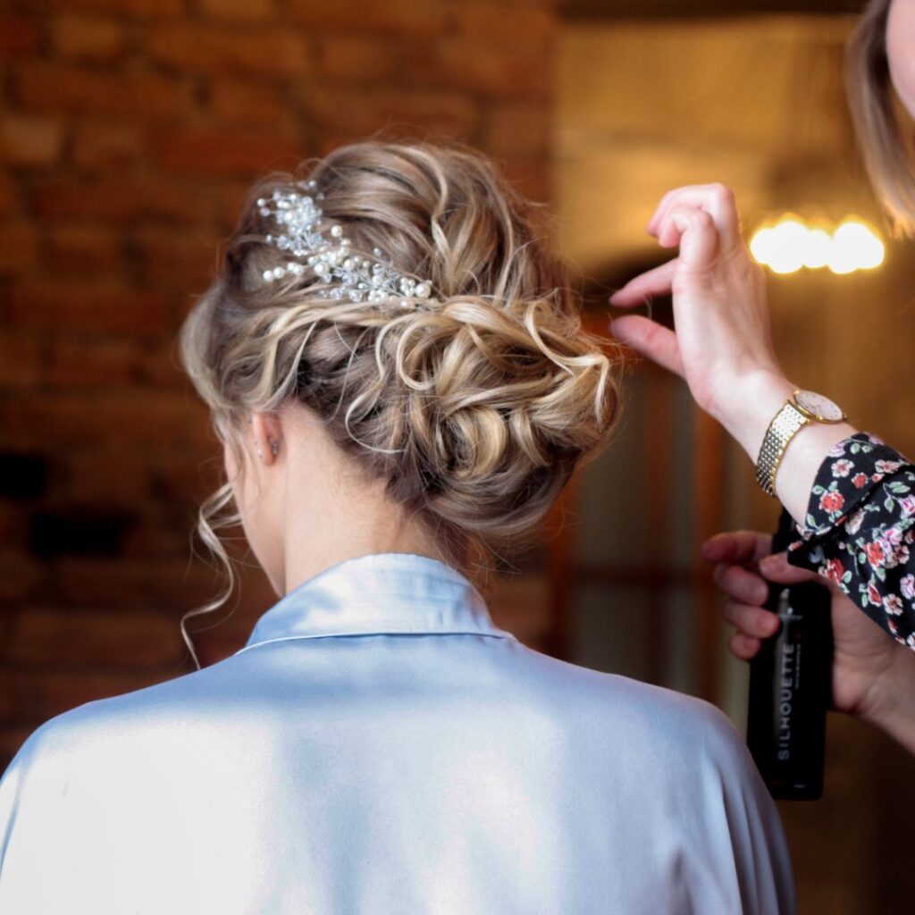 A person with styled blonde hair and a decorative hairpiece is having their hair adjusted by another person holding a spray bottle, capturing beautiful Wedding Hair Inspiration. - Detlev Lifestyle Salon in Coconut Grove, FL