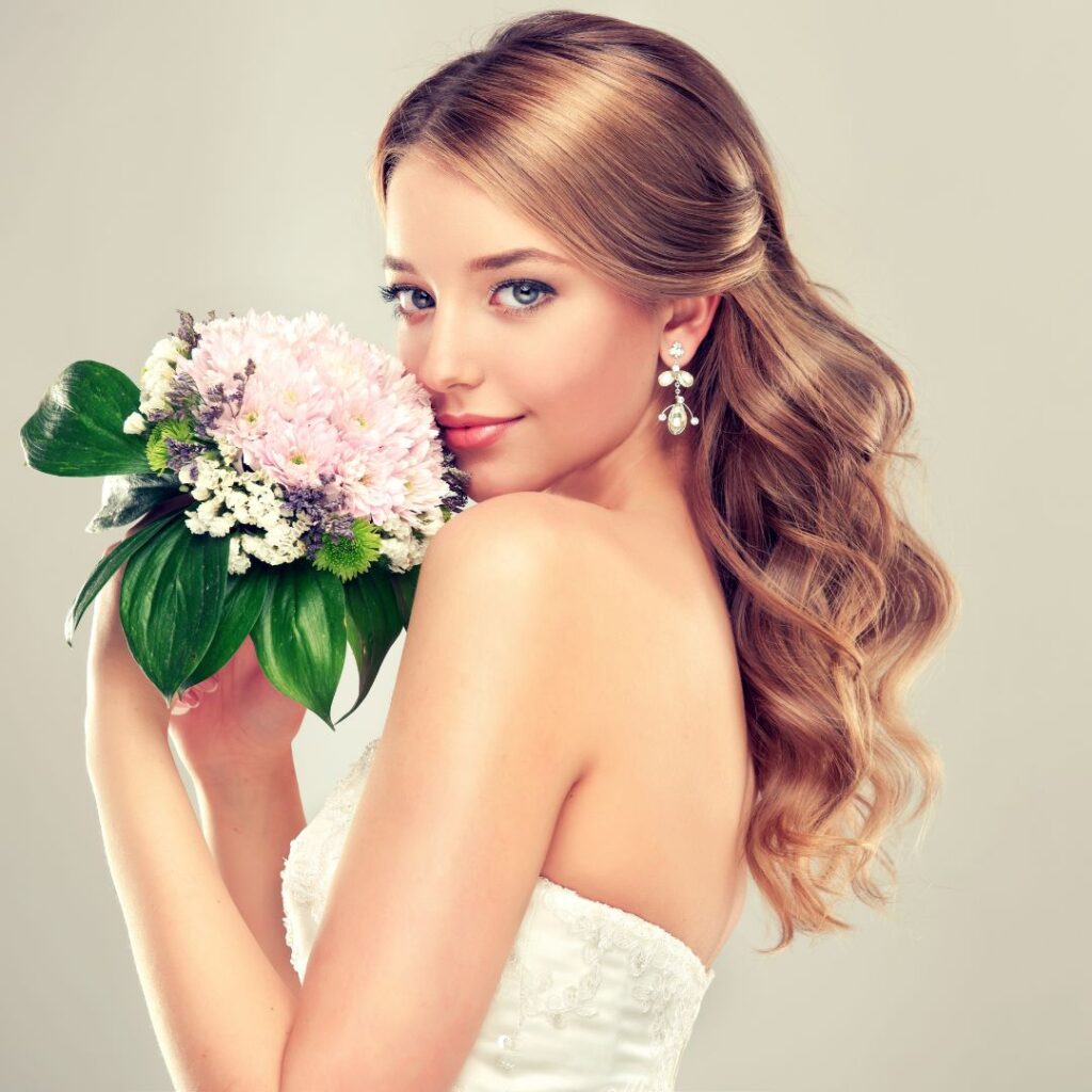 A woman in a white strapless dress holds a bouquet near her face, showcasing long wavy wedding hair and pearl drop earrings, posing elegantly against a plain background. - Detlev Lifestyle Salon in Coconut Grove, FL
