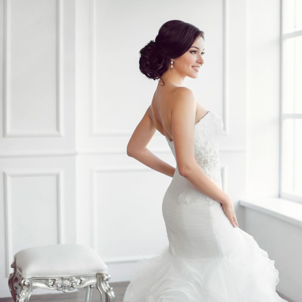 A woman in a strapless white wedding dress stands near a window, smiling and looking to the side; her elegant bridal hairstyle perfectly complements the look, while an ornate chair sits nearby. - Detlev Lifestyle Salon in Coconut Grove, FL