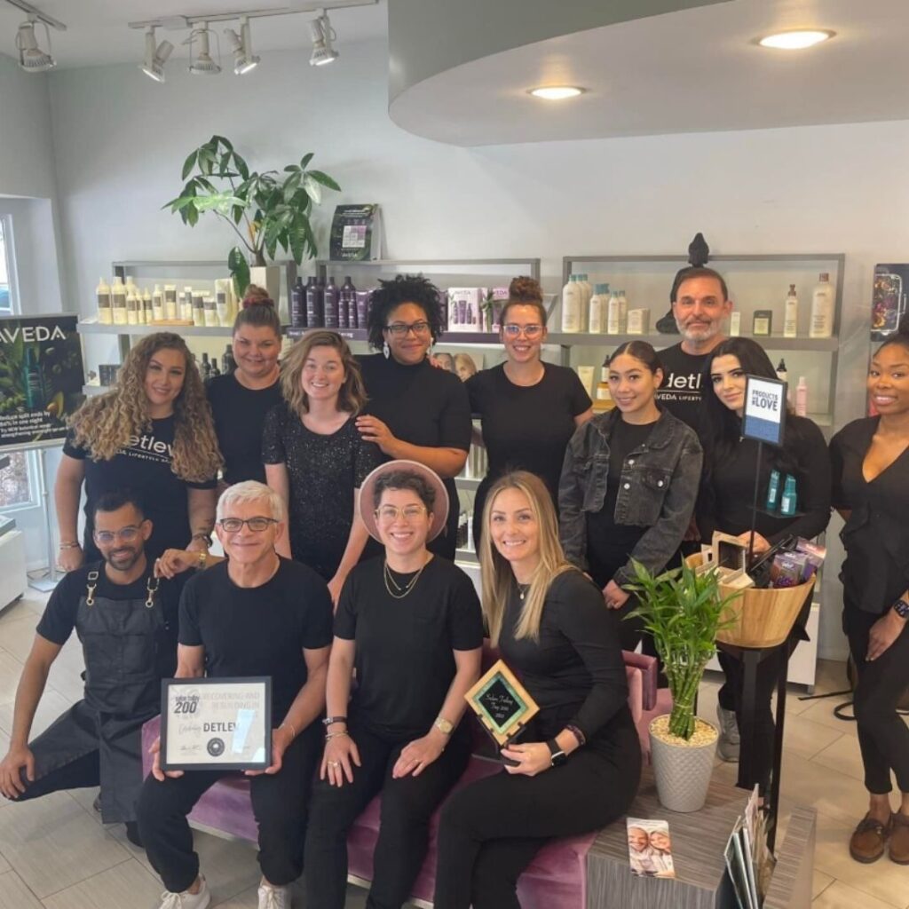 A group of 14 people in black clothing gather inside a salon, smiling and holding awards. Shelves with hair products and a plant add to the ambiance. It's the Detlev Difference that sets this celebration apart, highlighting excellence in every detail. - Detlev Lifestyle Salon in Coconut Grove, FL