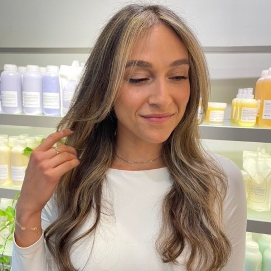 A person with long, wavy hair stands in front of shelves filled with hair products, embodying the Detlev Difference. Wearing a white top and touching their hair while looking down and smiling slightly, they capture the essence admired by salons everywhere. - Detlev Lifestyle Salon in Coconut Grove, FL