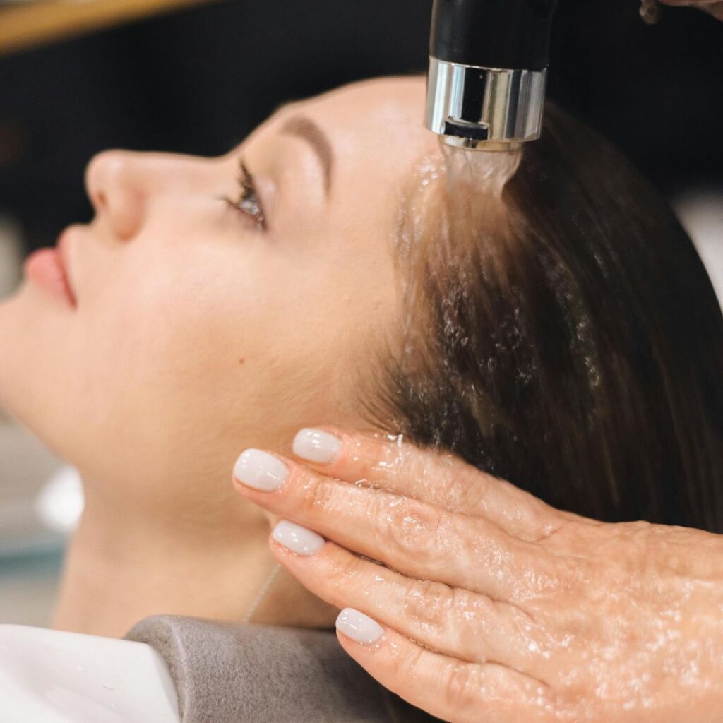 A person with long dark hair is having their hair washed at a salon sink, enjoying a unique salon experience as water flows from the faucet and a hand gently supports their head. - Detlev Lifestyle Salon in Coconut Grove, FL