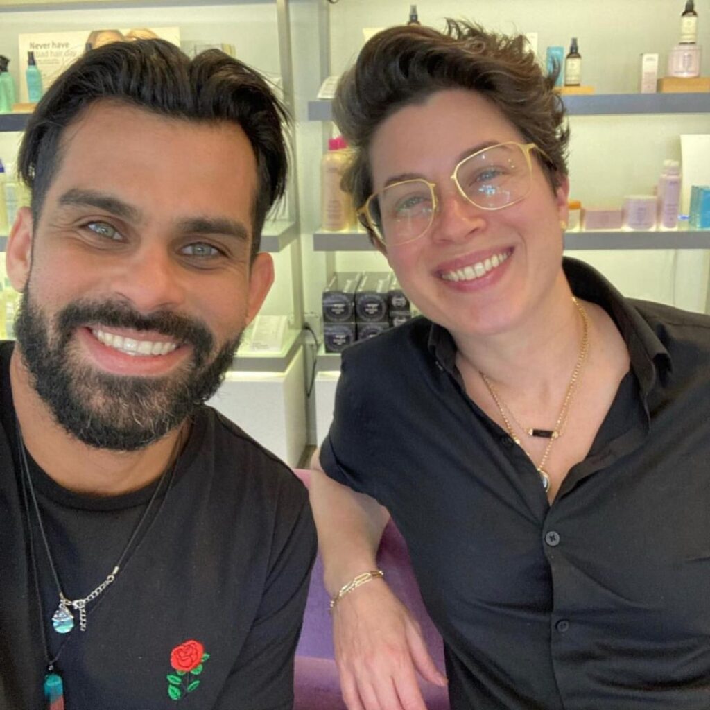 Two people are smiling at the camera inside a Coconut Grove hair salon, with shelves of skincare or beauty products in the background, capturing a unique salon experience. - Detlev Lifestyle Salon in Coconut Grove, FL