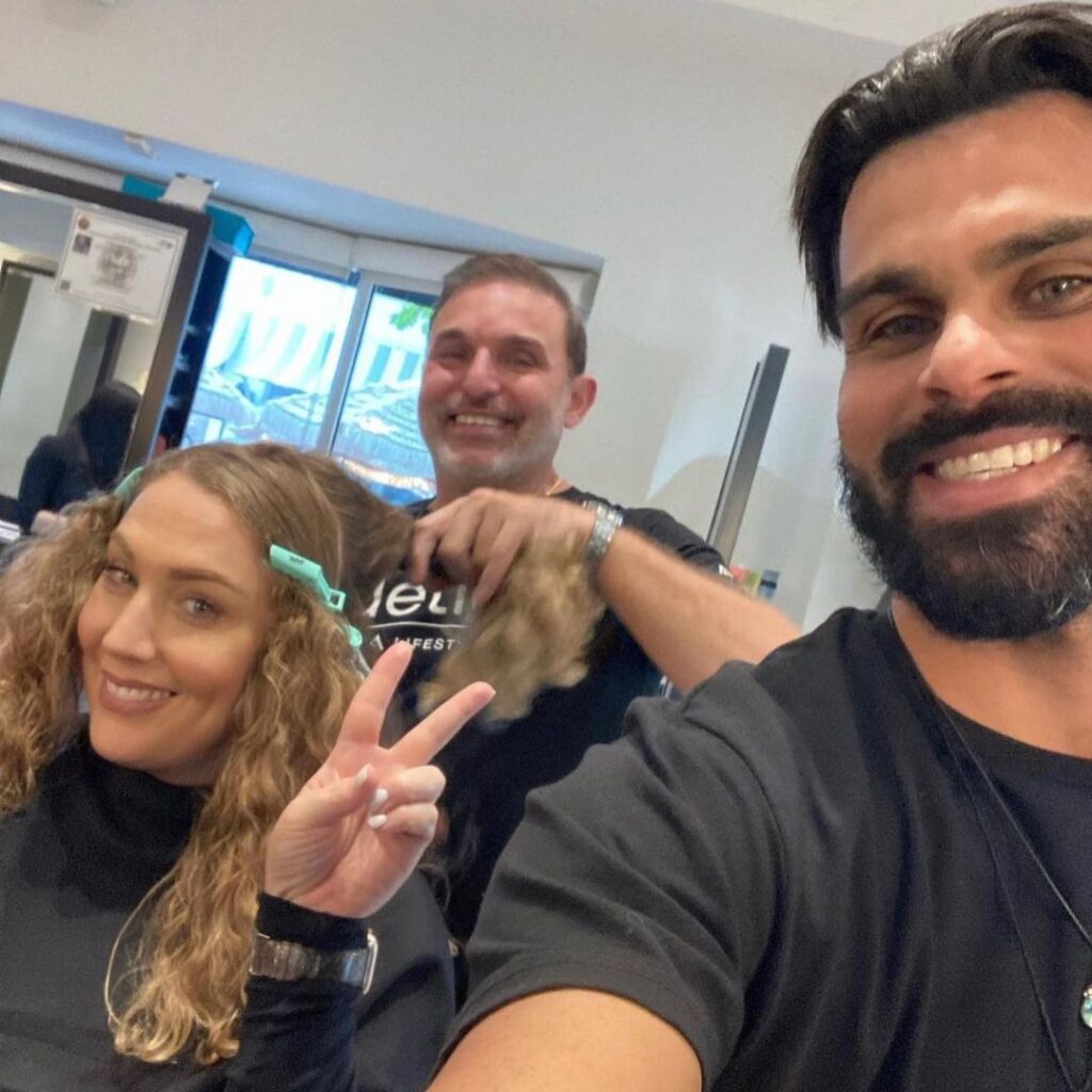 A woman with curly hair sits in a salon chair making a peace sign, while a stylist works on her hair and another person takes a selfie—capturing a unique salon experience at this Coconut Grove hair salon. - Detlev Lifestyle Salon in Coconut Grove, FL